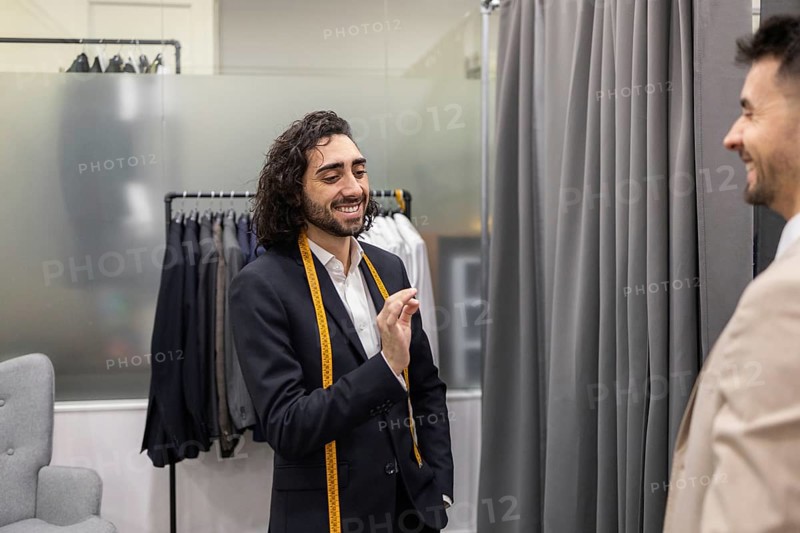 A tailor communicates with a customer in a suit store, showcasing a friendly ambiance. Suits hang in the background, highlighting the tailor expertise and the shop selection