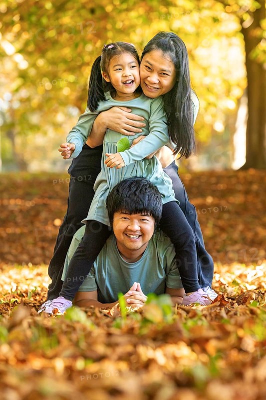 Asian family enjoying a playful moment outdoors in an autumn park. The child rides on the father's back, while the mother supports the child, surrounded by vibrant fall leaves