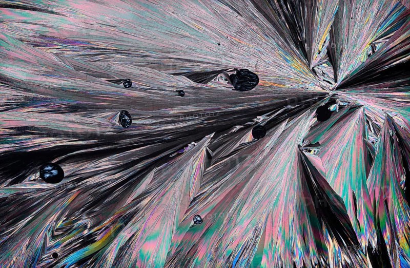 Stunning microscopic image showcasing the intricate patterns and vibrant colors of Hippuric acid crystals. Shades of pink, blue, green, and silver create a visually striking landscape