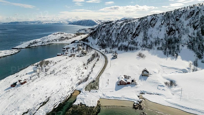 Aerial view of a serene Nordic landscape showcasing a snow-covered coastline, winding road, and scattered rustic houses against a backdrop of majestic snowy mountains, Botnhamn, Troms, Norway