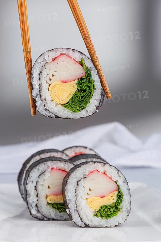 A close up view of sushi rolls with various colorful ingredients like crab, omelette, and greens wrapped in seaweed and rice in white background. Chopsticks hold one piece, showcasing its texture
