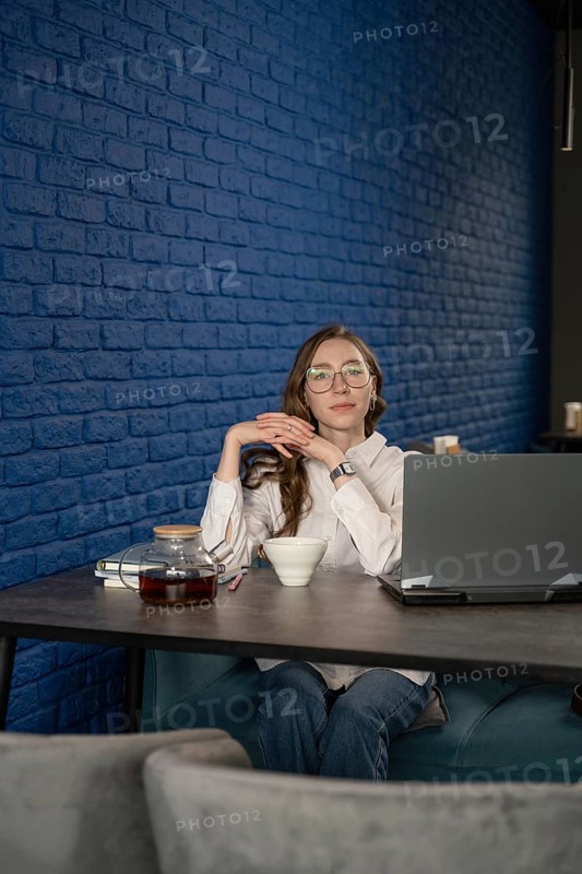 A woman with glasses sits at a cafe table with a laptop, tea cup, and teapot, against a stylish blue brick wall, taking a relaxing break from work