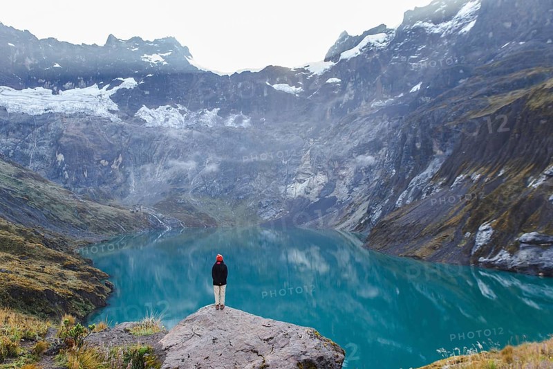 An Ecuadorian woman stands on a rocky ledge, overlooking the serene turquoise waters and majestic peaks of El Altar volcano in Ecuador, surrounded by misty clouds