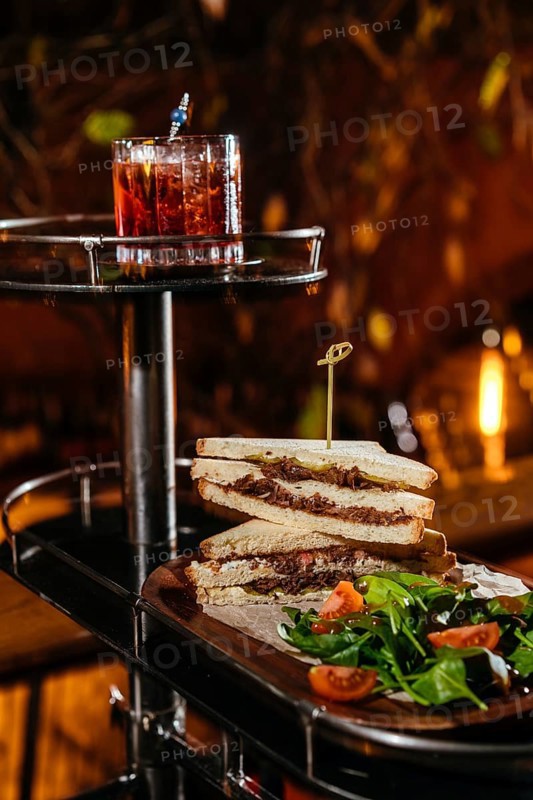 A gourmet setting features a stacked club sandwich with veal cheeks, cheese, and fresh greens, paired with a vibrant Negroni. Candlelight ambiance enhances the dining experience