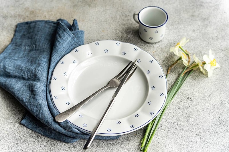 From above, a dining setup featuring a white plate with blue polka dots, accompanied by silverware on a blue napkin and a matching cup. Beside the plate, faded white daffodils add a touch of nature