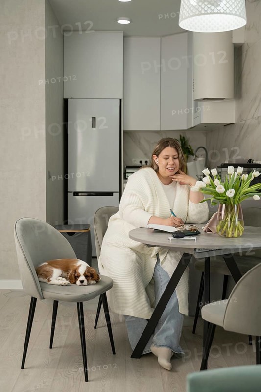 A woman sits at a modern kitchen table, writing in a notebook. Nearby, a small dog rests on a chair. Tulip vase adds elegance to the cozy, light-filled space