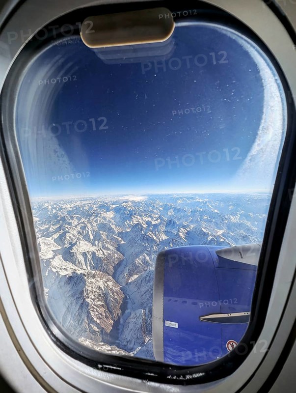 Stunning aerial view of a vast snow-covered mountain range as seen through an airplane window. The image captures the serene beauty of the landscape from high above the clouds