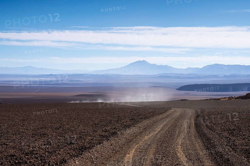 A dusty, gravel road cuts through the arid landscape of La Puna in Argentina, offering a view of distant mountains under a clear blue sky
