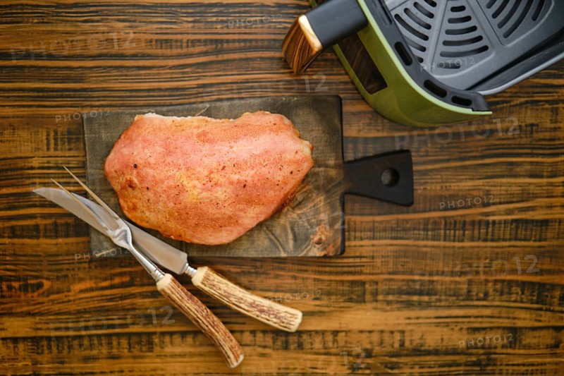 A piece of roasted pork meat on a wooden cutting board. Kitchen knife and fork are placed next to the fillet. An air fryer is seen on the side of the board. Photo with shallow depth of field