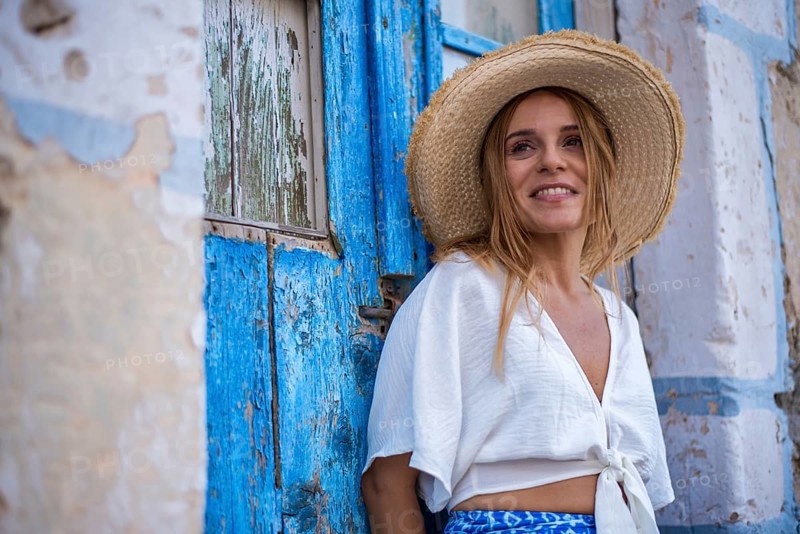 A smiling woman in a straw hat leans against a rustic blue door in Trypiti, Milos Island, Greece. The setting exudes a relaxed summer vibe with a touch of Mediterranean charm