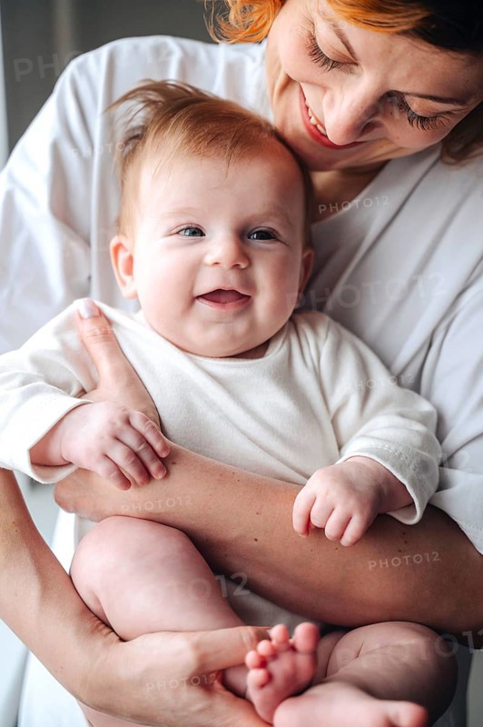Loving mother cradles smiling newborn baby girl at home, both wearing white clothes. The baby's joyful expression enhances the warmth of their shared moment