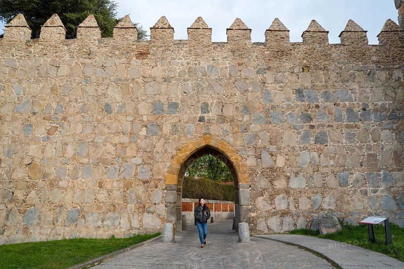 An Asian woman walks through an arched entrance of a medieval city wall in Avila, Spain, surrounded by historic stone architecture, showcasing the blend of history and culture