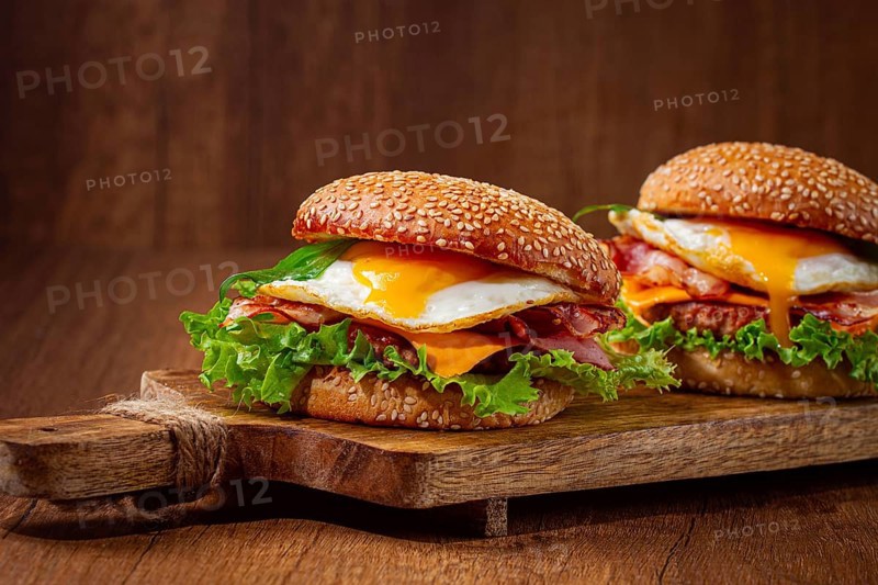 Two juicy burgers sit on a wooden board, each filled with lettuce, bacon, cheese, and fried eggs