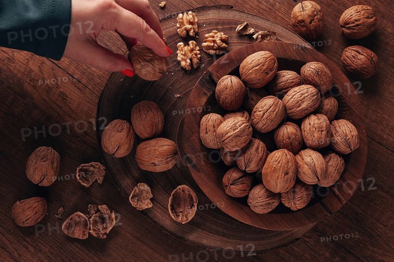 A woman's hand taking a walnut from a wooden bowl filled with walnuts. There are shells and walnuts scattered around