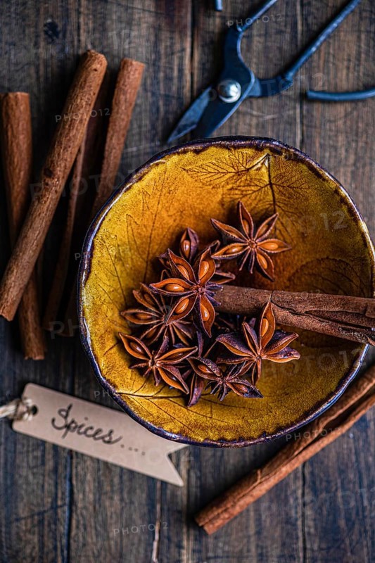 Top view of a rustic yellow bowl filled with star anise and cinnamon sticks, accompanied by a pair of old scissors and a label reading Spices. Perfect for illustrating cozy, aromatic culinary themes