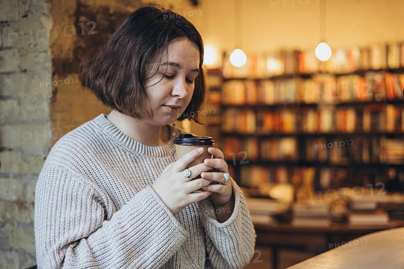 A woman in a soft sweater holds a takeaway coffee cup while enjoying a peaceful moment in a cozy bookstore Bookshelves in the background create a warm and relaxed atmosphere