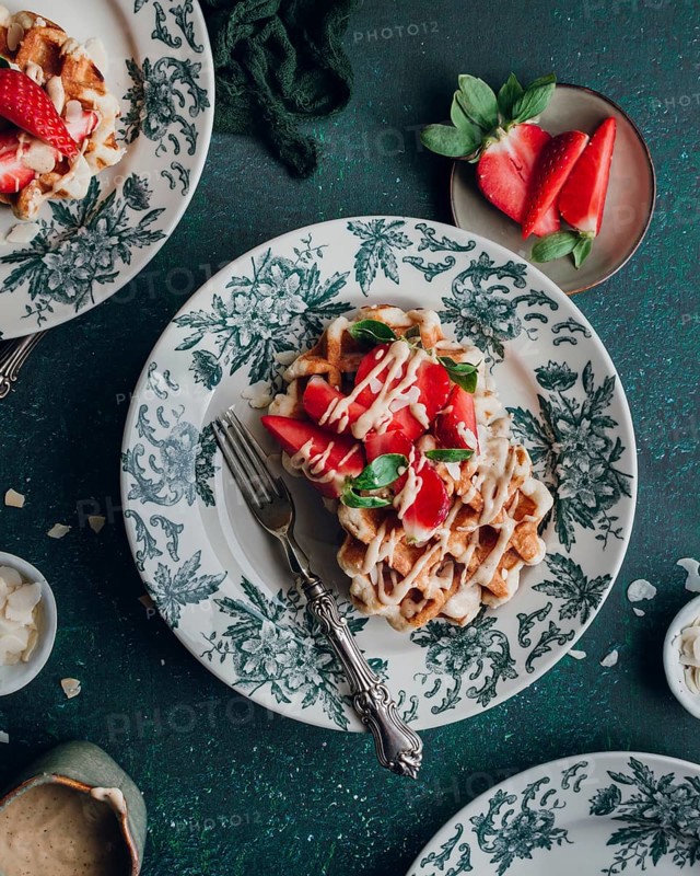 Delicious waffles topped with fresh strawberries and cream are elegantly served on beautiful floral-patterned plates, creating a visually appealing and mouth-watering breakfast scene