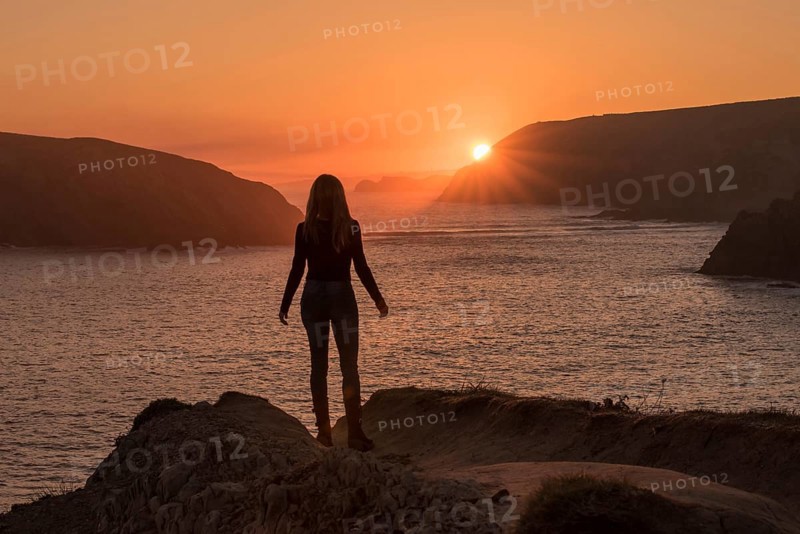A silhouette of a woman stands on a rocky cliff at Arnia Beach, Costa Quebrada, gazing toward the glowing sunrise over the Atlantic coastline in northern Spain