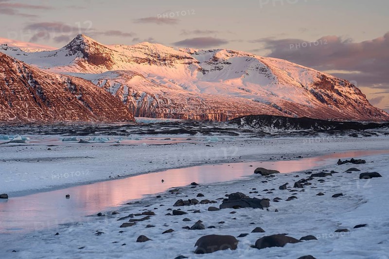 Majestic Icelandic landscape with sunset hues on snow-covered peaks, reflected in icy waters. A stunning display of nature in Iceland's winter wonderland setting in Skaftafell