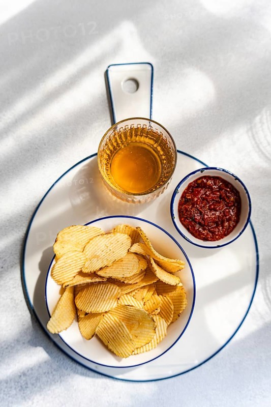 A plate of homemade potato chips served with a small dish of red sauce and a glass of golden beverage. The setup is on a white surface with natural lighting