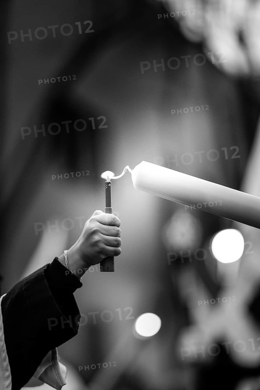 A monochrome image depicting a hand holding a lighter, reaching to ignite a tall candle, symbolizing light and spirituality in holy week. The blurred background enhances the focus on the flame