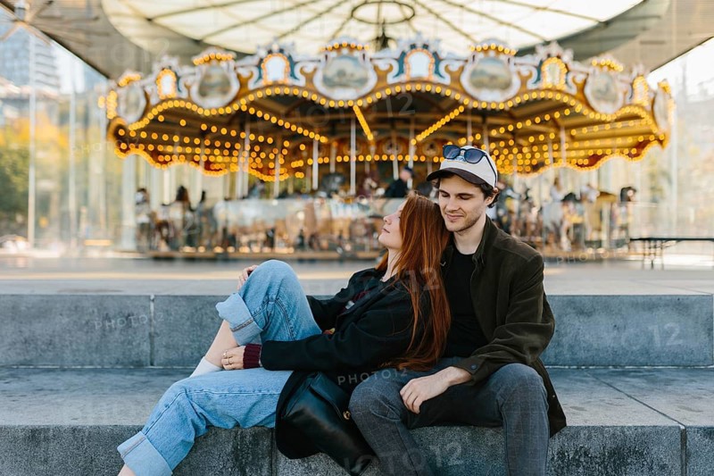 A young couple sits on steps in front of a brightly lit carousel in Brooklyn Bridge Park. They exude relaxation and happiness, enjoying a sunny day in an urban setting, surrounded by street activity