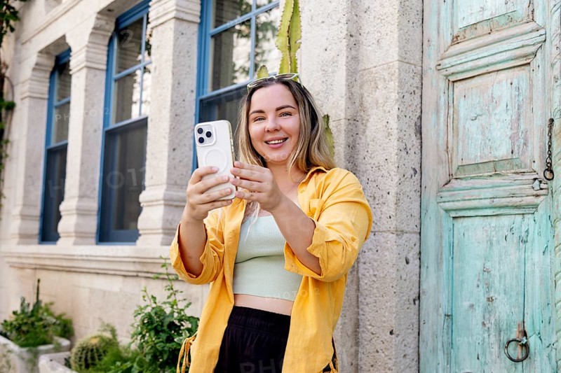 A cheerful female tourist wearing a yellow shirt uses her smartphone to capture memorable moments while traveling, standing in front of a historical building with blue windows