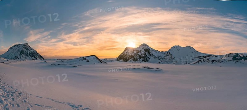 The sun sets behind the rugged, snow-covered peaks of Katla Glacier, casting a golden glow over the vast, untouched winter landscape. A dramatic sky completes this breathtaking panoramic view