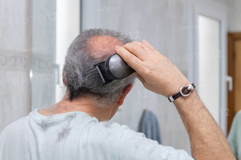A mature man uses an electric clipper for a self-haircut, highlighting the trend of personal grooming at home. The image emphasizes simplicity and self-care