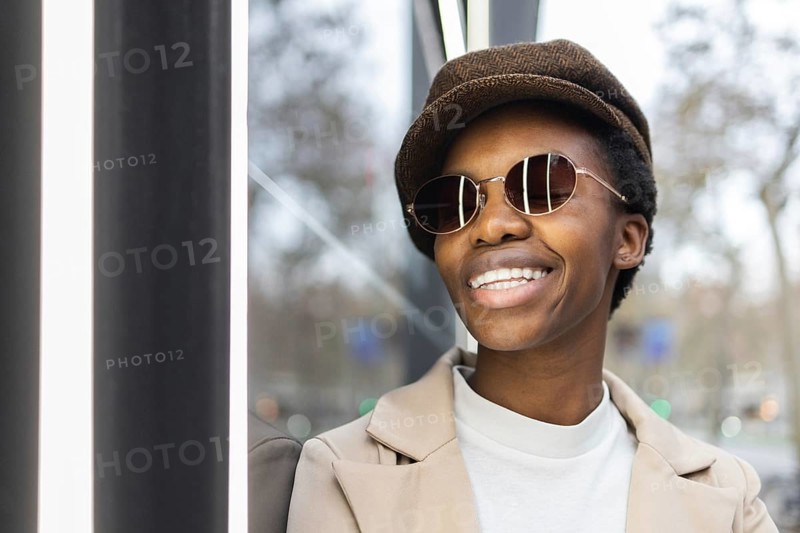 A fashionable black woman wearing sunglasses and a hat, smiling confidently in an urban setting Her stylish outfit and accessories highlight her vibrant personality
