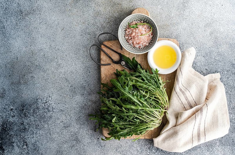 Fresh arugula, Himalayan pink salt, and olive oil arranged on a rustic wooden cutting board with vintage scissors and a linen towel, styled for a natural kitchen aesthetic