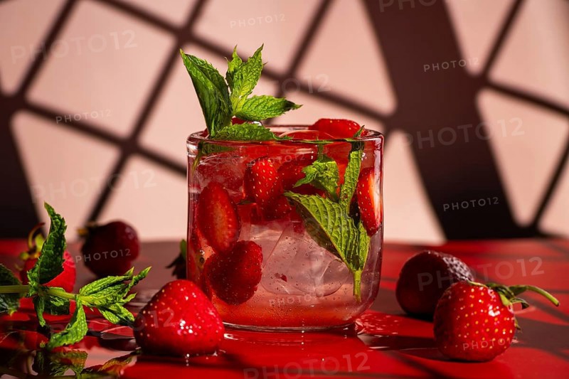 A refreshing strawberry mojito with fresh strawberries and mint, served in a clear glass with ice. On a red surface against a pink background, enhancing the cocktail's vivid colors