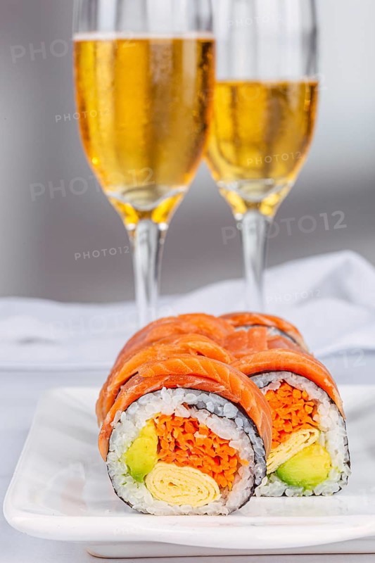 Delicious sushi rolls with salmon, avocado, and omelette are elegantly displayed on a plate, complemented by two sparkling wines in the white background, creating a gourmet dining scene