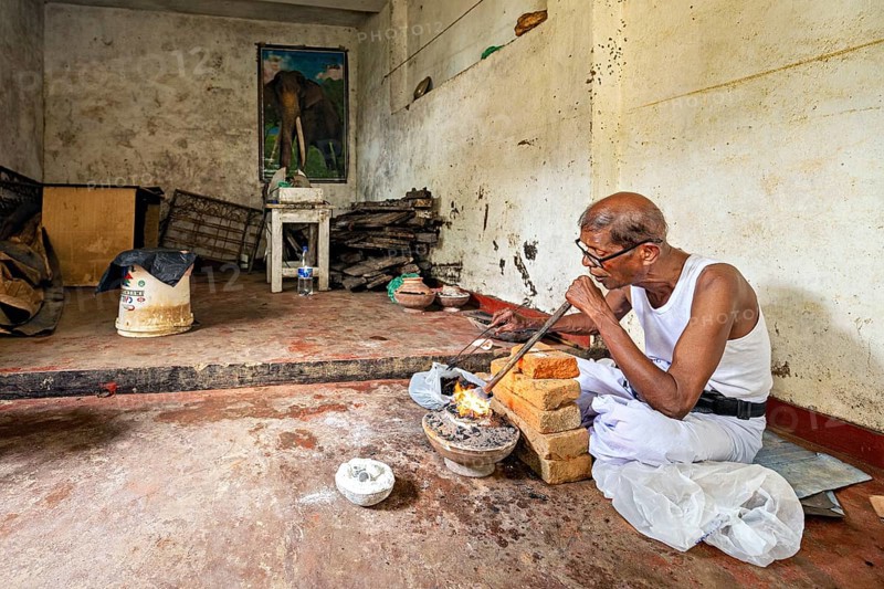 An old man works silently in a workshop with bricks and fire, an elephant poster on the wall, a man burns sapphires in a small oven in the town of Ratnapura in Sri Lanka