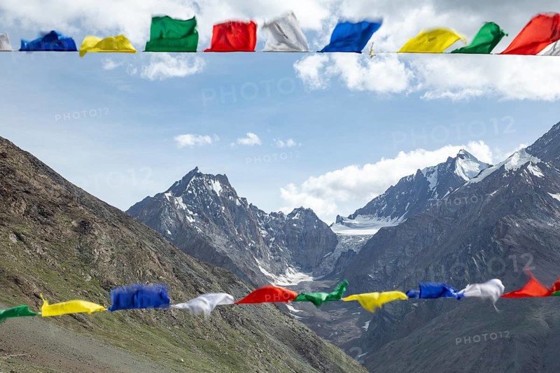 Colorful prayer flags flutter in the foreground, set against the breathtaking backdrop of the majestic mountains in Spiti Valley, India, under a bright blue sky