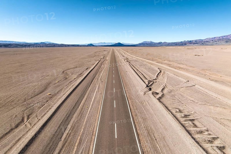 A straight road stretches across a vast desert landscape of the argentine Puna, leading into distant mountains under a clear blue sky, evoking a sense of freedom and adventure