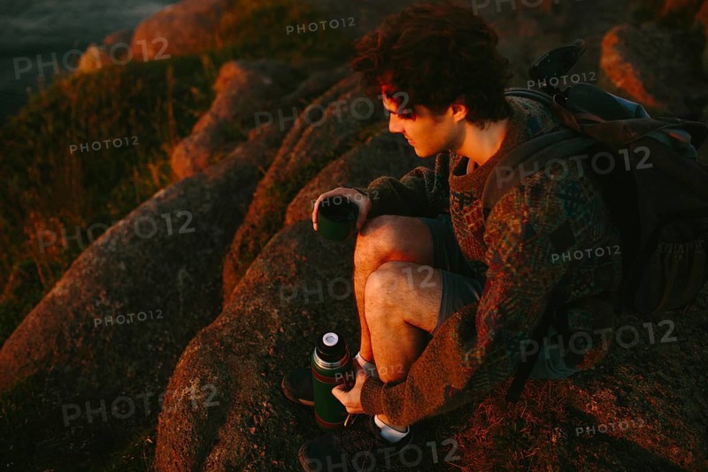 A young male hiker sits on rugged rocks at Cape Spear Lighthouse, watching the sunset. He is holding a thermos and relaxing after a day of hiking