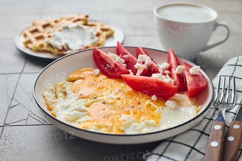 From above a delicious homemade breakfast featuring fried eggs with feta cheese and fresh tomato slices alongside Belgian waffles topped with cream cheese and a steaming cup of coffee