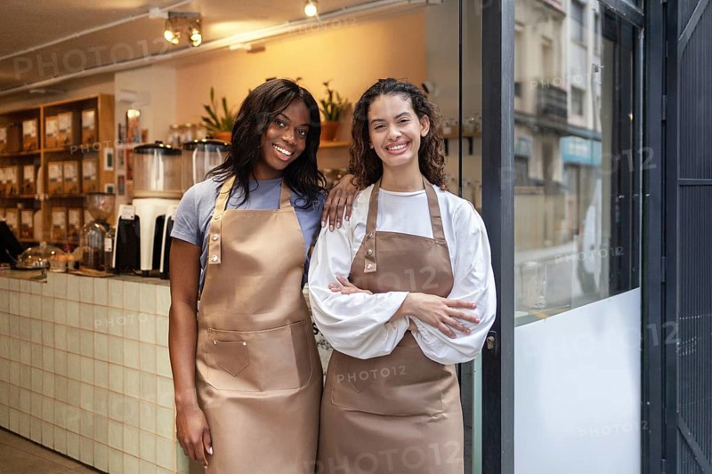 Two friendly clerks stand proudly at the entrance of an organic bulk store, wearing aprons and smiling. The store offers a variety of natural products for eco-conscious shoppers