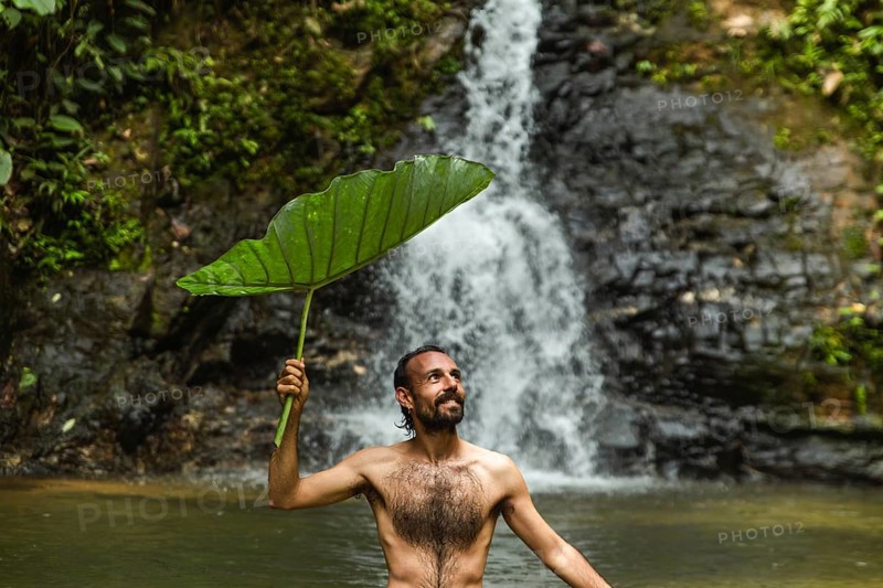 Shirtless man holding a large leaf smiles as he uses it for shade while standing in a water pool in front of a waterfall in the lush Choco Andino rainforest of Ecuador