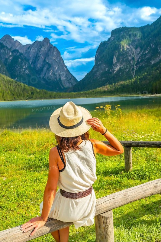 Woman wearing straw hat admiring lake dobbiaco with the dolomites in the background on a sunny summer day