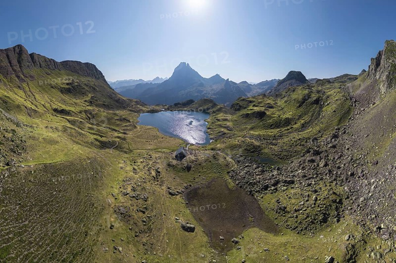 A stunning aerial view captures the tranquil Ayous Refuge surrounded by lush green landscapes and a pristine lake in the heart of the French Pyrenees under a clear blue sky