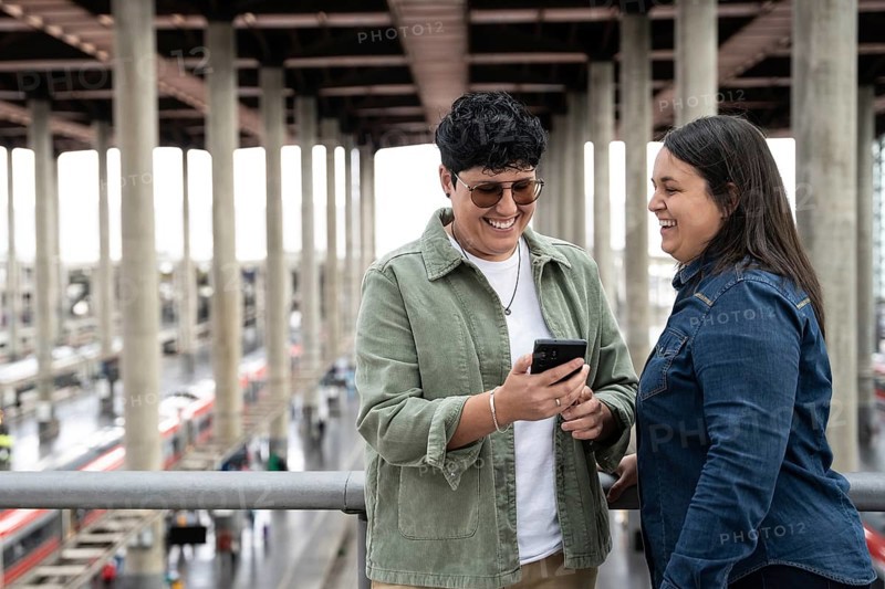 A joyful moment between a lesbian curvy couple at a train station, sharing smiles and a phone, embodying love and companionship in a travel setting