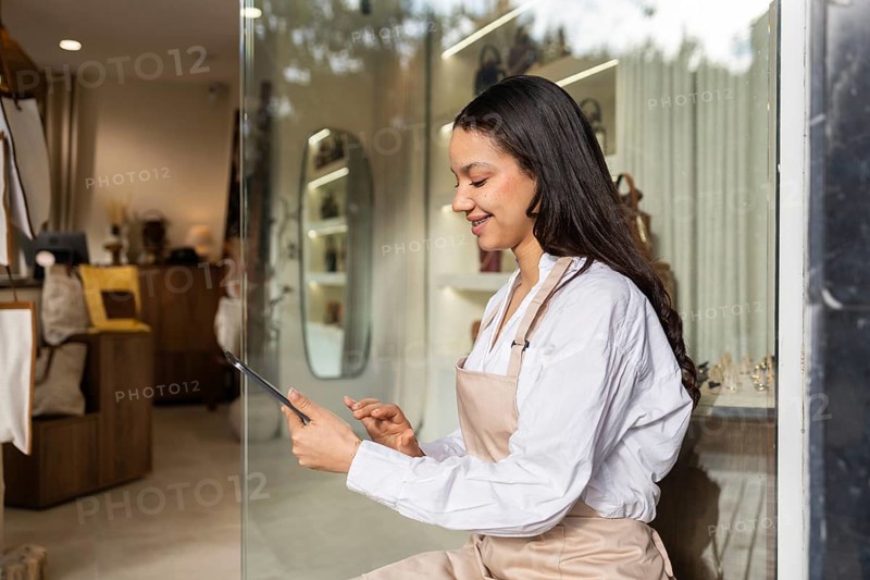 A young shop assistant in a modern bag store uses a tablet. She is standing by a glass display, surrounded by various stylish bags and accessories