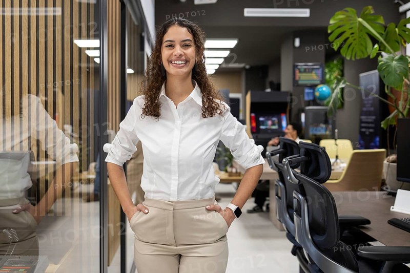 A woman in a white blouse stands confidently in a modern office. Her smile reflects a positive and professional demeanor, with stylish decor and workspace ambiance
