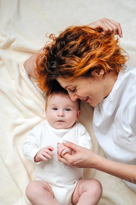 A mother shares a tender moment with her newborn baby girl, lying on a soft, cream-colored blanket. The baby wears a white onesie, creating a serene and loving atmosphere