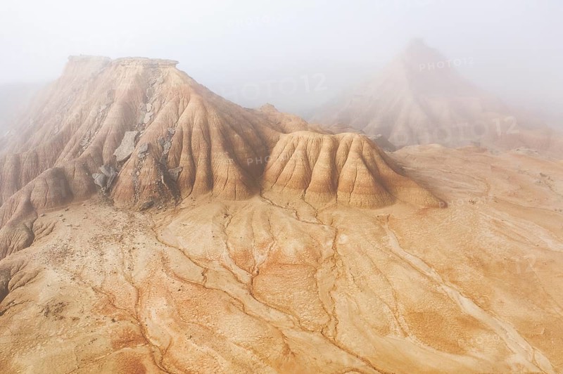 Aerial view of rugged desert formations enveloped in mist Erosion patterns are visible on the sandy surface, creating a mysterious and surreal atmosphere