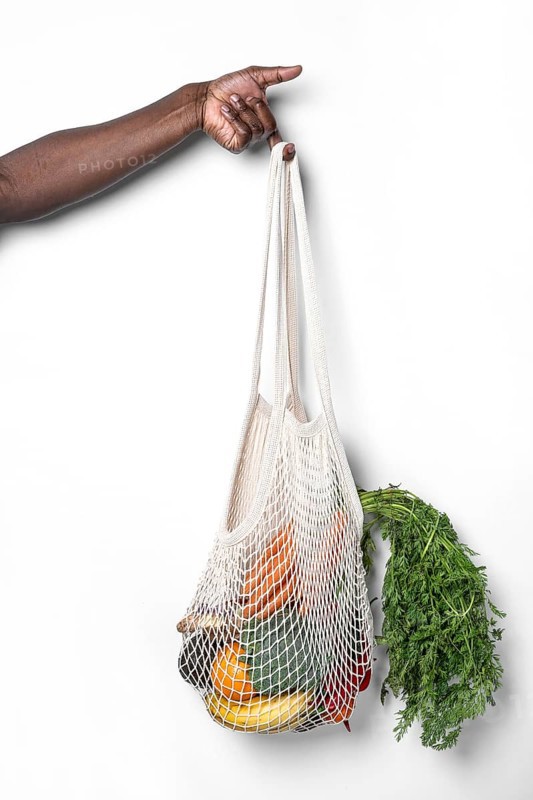 A hand holds a reusable cotton mesh bag filled with fresh vegetables and fruits, showcasing sustainable shopping practices against a clean white background, promoting eco-friendly habits