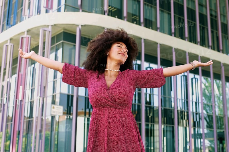 Young Arab woman with afro hair, wearing a pink dress, joyfully extends her arms in front of a modern glass building with eyes closed. She exudes confidence and empowerment
