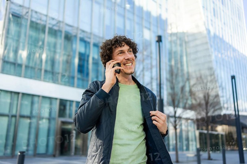 A businessman stands outside a sleek, modern glass building, smiling as he talks on his smartphone, radiating positivity and connection in a bustling urban environment